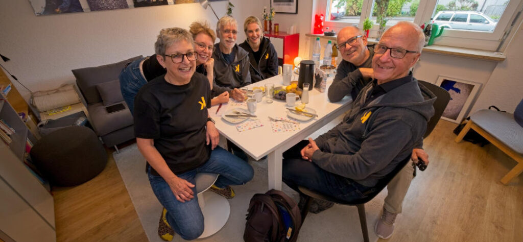 Fünf Personen sitzen lachend um einen Tisch in einem hellen Raum. Auf dem Tisch stehen Tassen, Teller und eine Kaffeekanne; zwei Personen tragen schwarze Oberteile mit gelbem Logo.